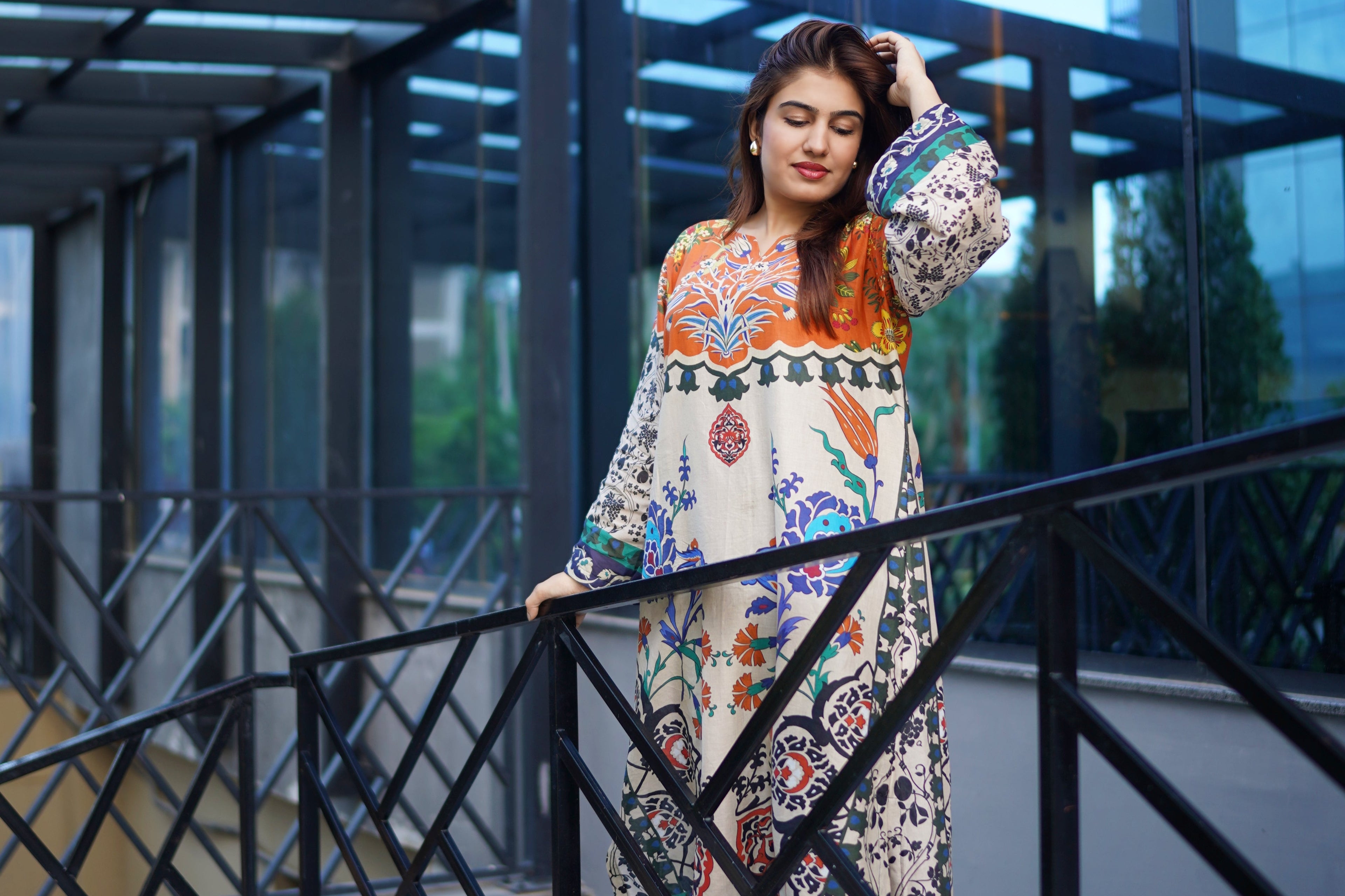 A woman posing on outdoor stairs in front of a modern glass building, wearing a long-sleeved cream-colored printed kurta with orange, green, and blue patterns, paired with black pants.